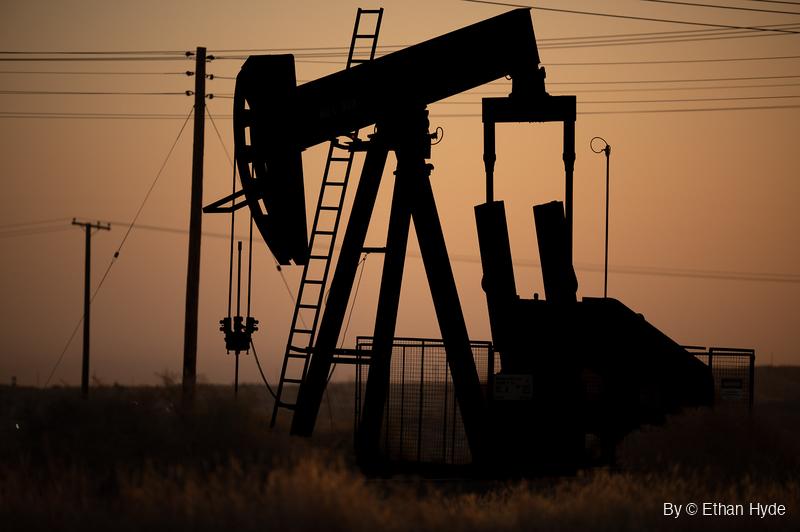 A close silhouette of an oil pumpjack with its ladder and counterweights against an orange sunrise sky, power lines and poles behind.