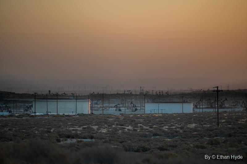 Rows of pumpjacks and storage tanks fade into amber haze across a flat at sunrise.