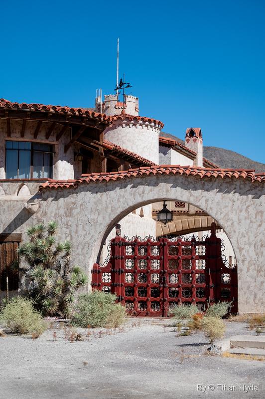 Wood and wrought-iron courtyard gates at Scotty's Castle under a stucco arch, with a round tower and weathervane above.