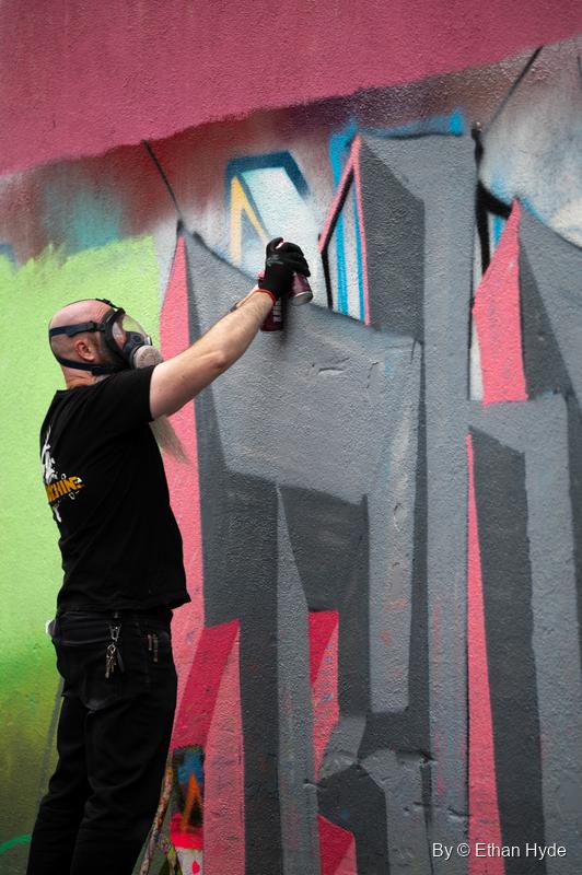 An artist in a respirator mask sprays gray and pink block letters on a wall.