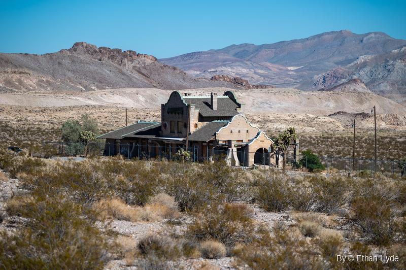 A mission-style block railroad depot with curved gables and a dark shingle roof stands behind a chain-link fence in desert scrub below bare hills.