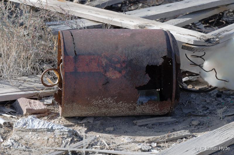 A rusted metal can with a ring handle lies on its side among weathered gray boards, coiled wire and dry brush, a glass bottle visible through a hole.