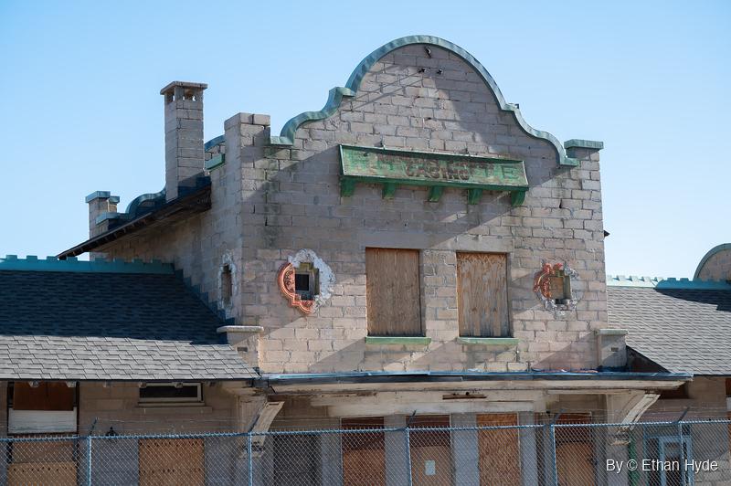 The scalloped parapet of an old concrete-block railroad depot with two boarded upper windows and green trim, above a chain-link fence.