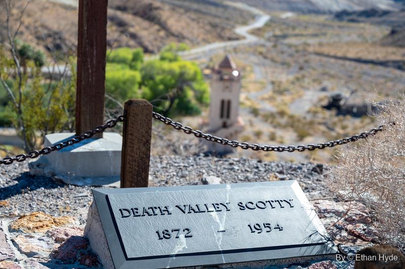A grave marker reading "Death Valley Scotty 1872–1954" on Windy Hill, with a chain fence and wooden cross and the Chimes Tower below.