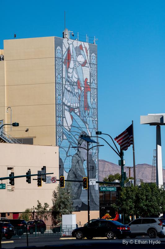 A mural in a stained-glass style covers a tall building wall above traffic lights, an American flag and desert mountains.