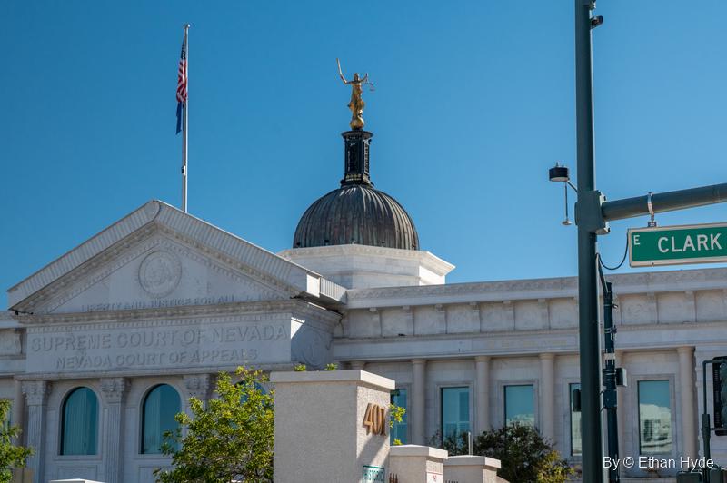 The white Nevada Supreme Court building with its domed cupola, Lady Justice statue and flag behind a green Clark Avenue street sign.