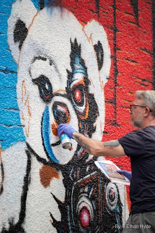 A gray-haired artist in blue gloves sprays a mural of a teddy bear holding a camera against a red brick-pattern background.