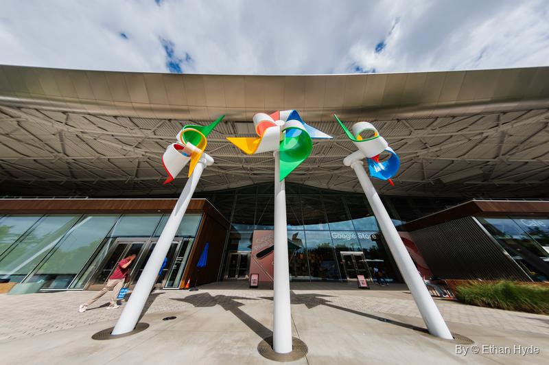 Three red, green, yellow and blue pinwheels on leaning white poles in front of Gradient Canopy's glass entrance.