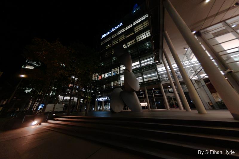 Stacked white disc forms of a large sculpture stand before a lit glass office building and wide steps at night.