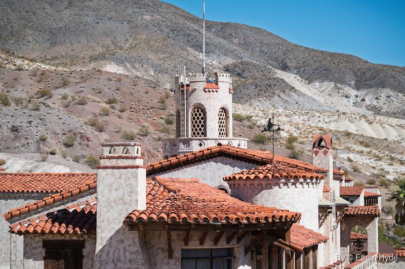 Red tile roofs and chimneys of Scotty's Castle beneath a round crenellated tower and a weathervane, with banded desert hills behind.