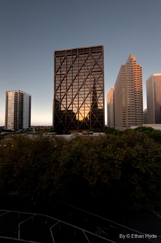 A dark grid-faced office tower catches orange dusk light between pale high-rises, above a band of treetops.