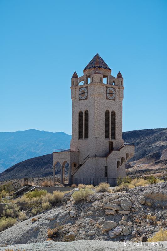 The Chimes Tower at Scotty's Castle seen from below on a rocky slope, with distant desert mountains under a clear sky.
