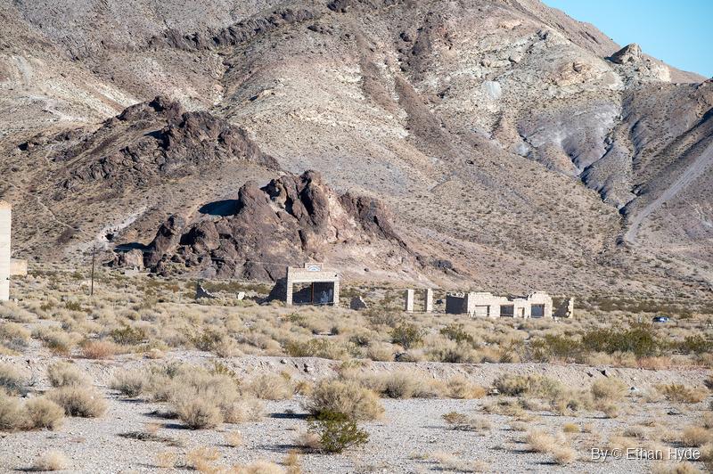 Roofless stone building shells and a freestanding facade stand in dry brush beneath a dark rock outcrop and a steep desert mountainside.
