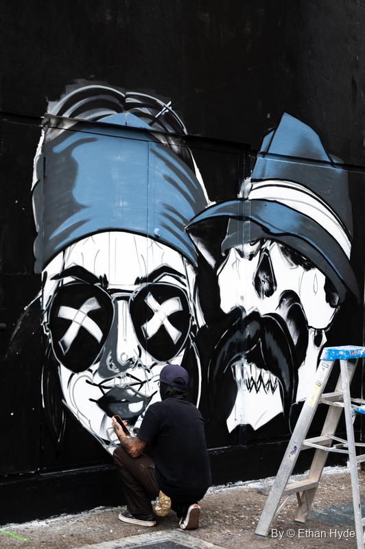 An artist crouches to paint a black-and-white mural of two skull-faced figures in caps, one wearing sunglasses with X's for eyes.