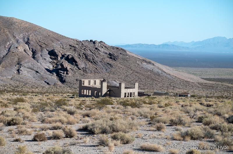 A roofless two-story concrete ruin with empty window openings stands in desert scrub below a rocky hill, with a wide basin and hazy mountains beyond.