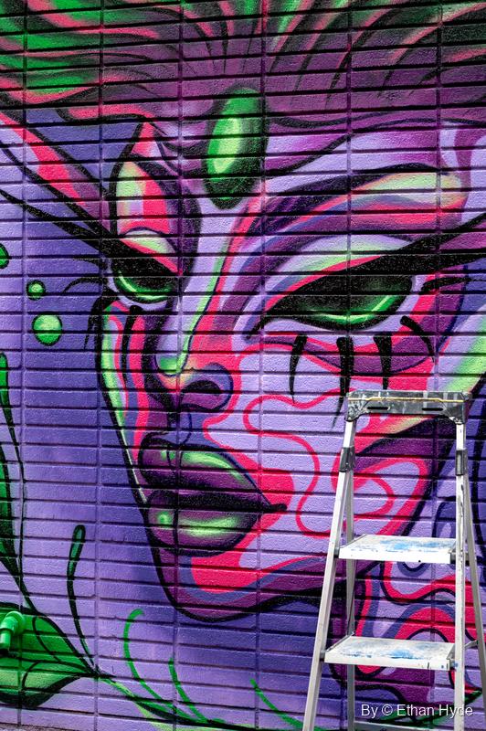 A purple, pink and green face with glowing green eyes painted across a roll-up metal shutter, a stepladder in front.