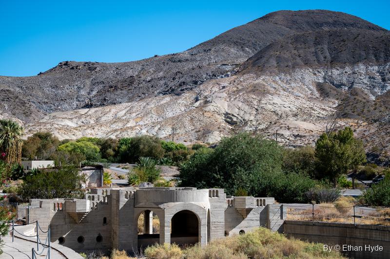 Crenellated walls and arched openings around the unfinished pool area at Scotty's Castle, with oasis trees and banded hills behind.