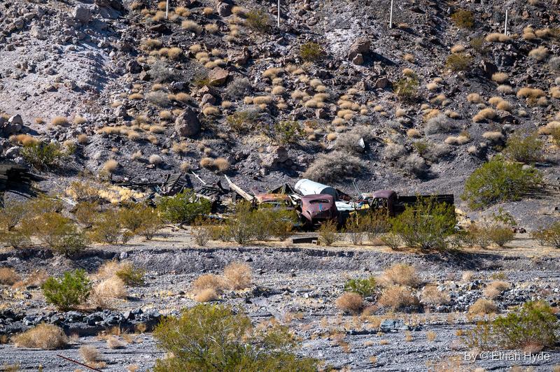 Rusted vintage car bodies and debris in a desert wash below a scrub-covered hillside near Scotty's Castle.