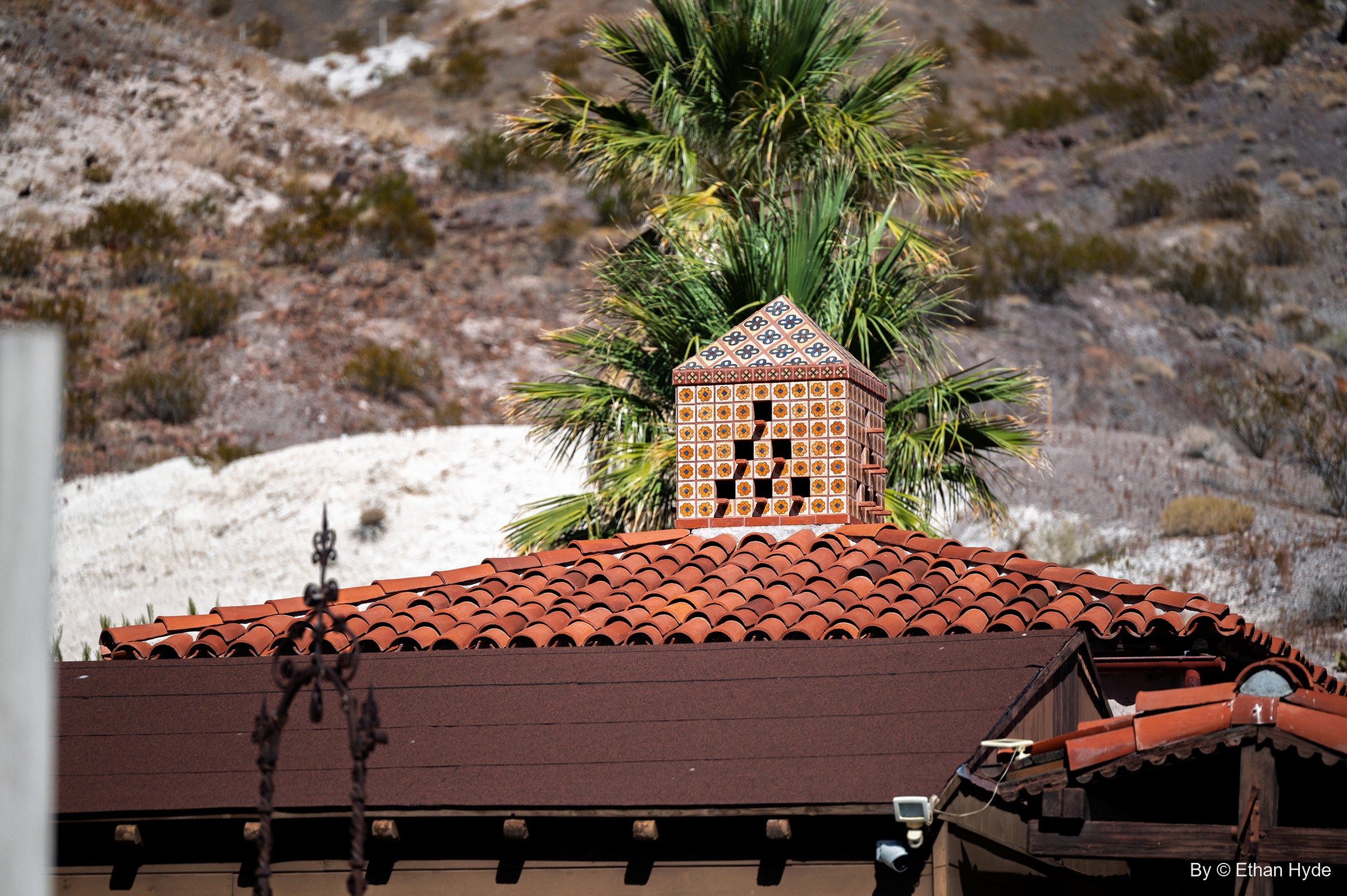 A patterned tile chimney cap on a red tile roof at Scotty's Castle, a palm and desert hillside behind.