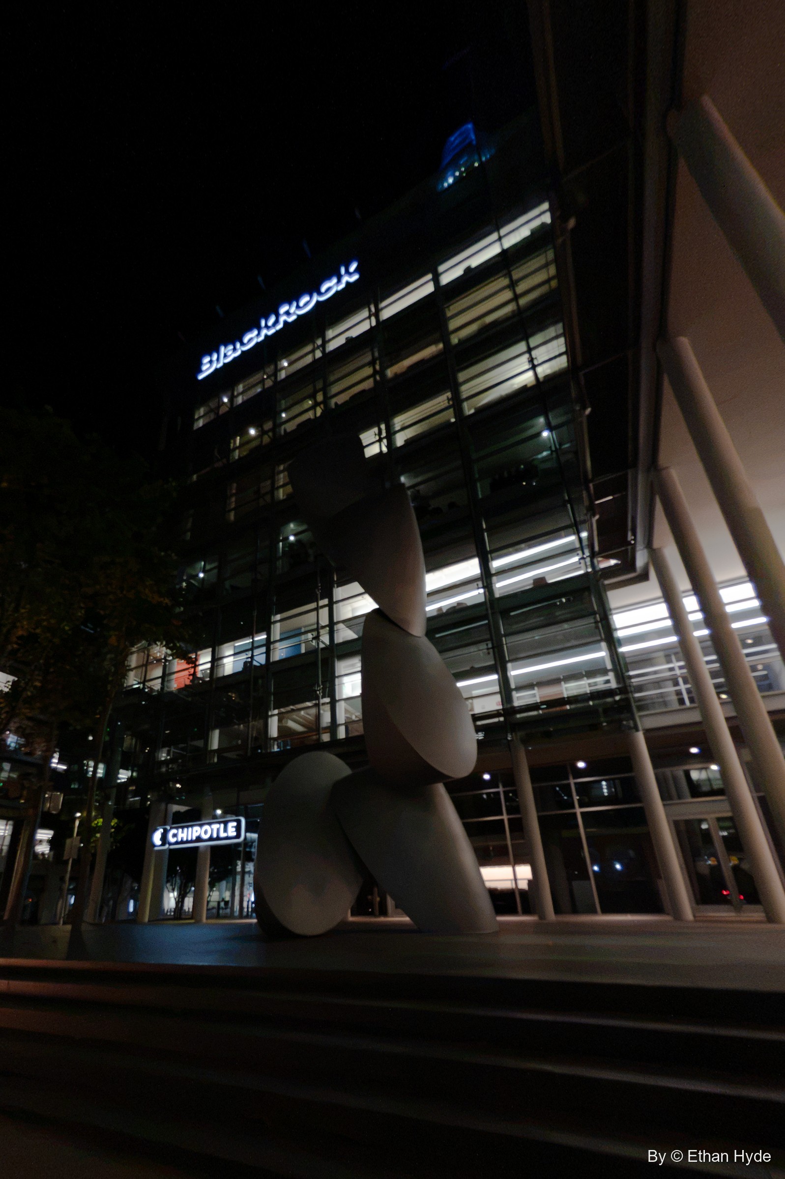 A tall sculpture of stacked white disc forms rises in front of a glass office building with a lit BlackRock sign at night.