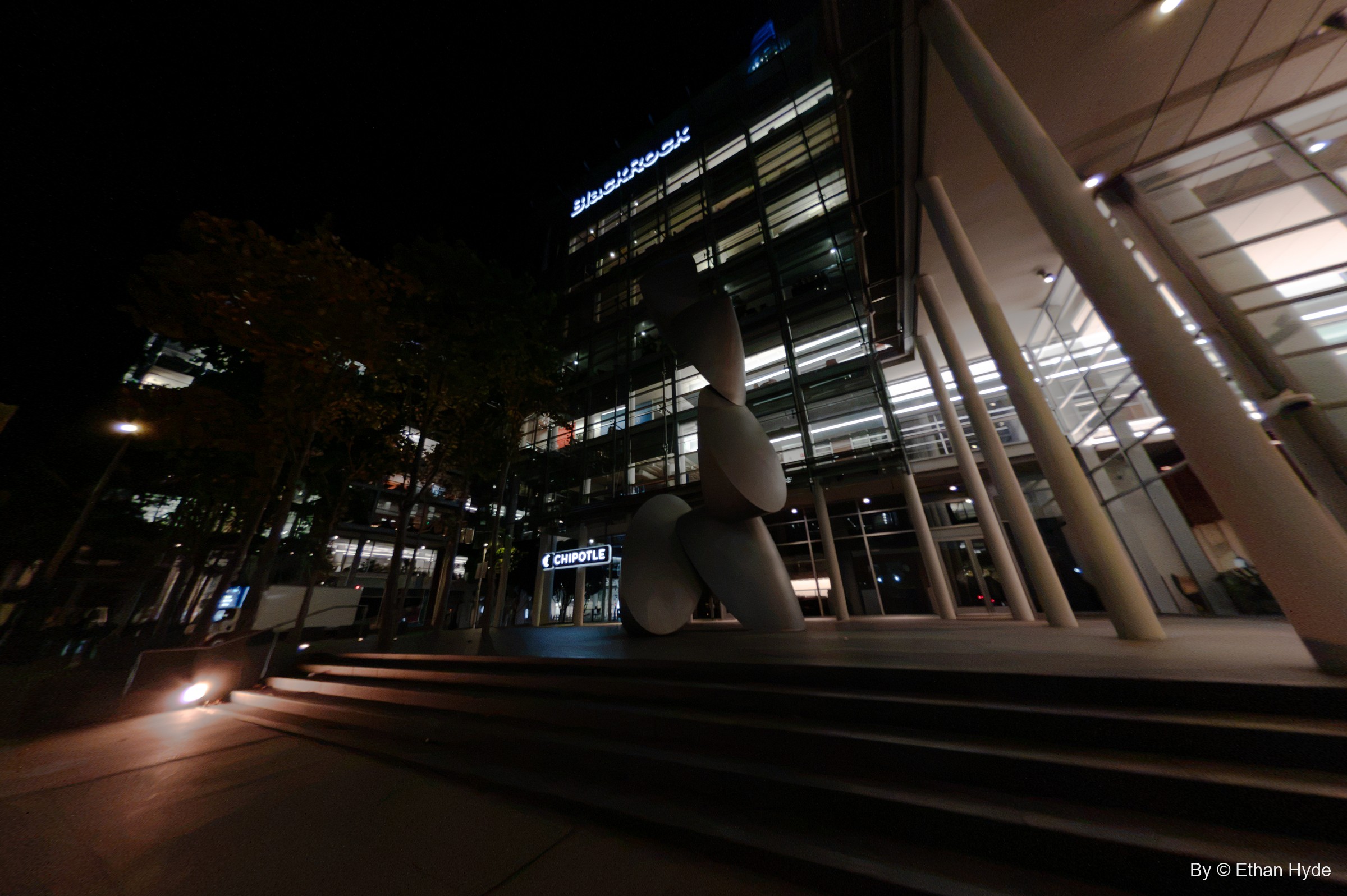 Stacked white disc forms of a large sculpture stand before a lit glass office building and wide steps at night.