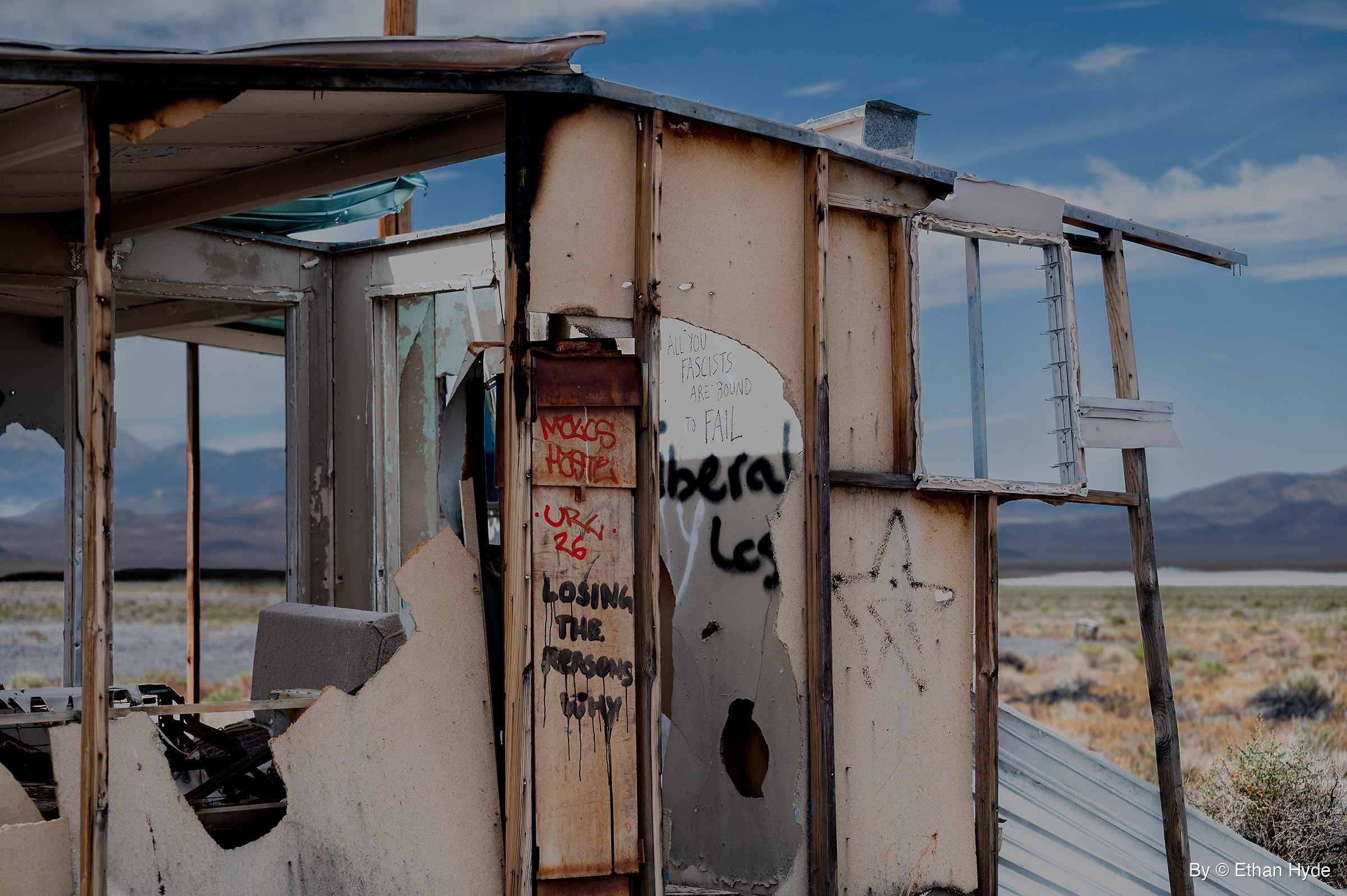 A roofless desert shack with torn plywood walls covered in spray-painted slogans, with mountains and a pale dry lake bed behind it under a blue sky.