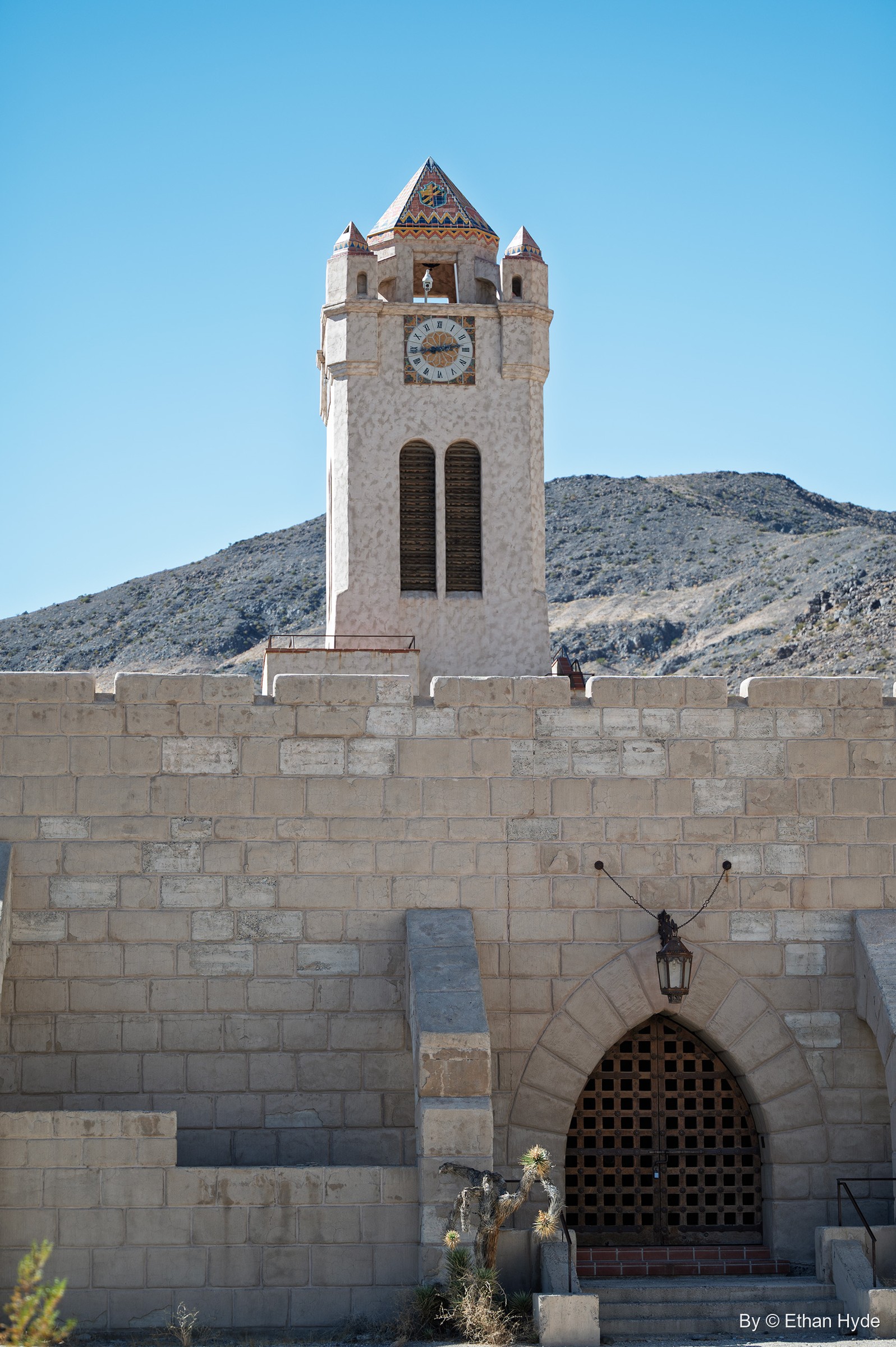 The Chimes Tower at Scotty's Castle rises behind a crenellated block wall with a pointed-arch iron gate and lantern.