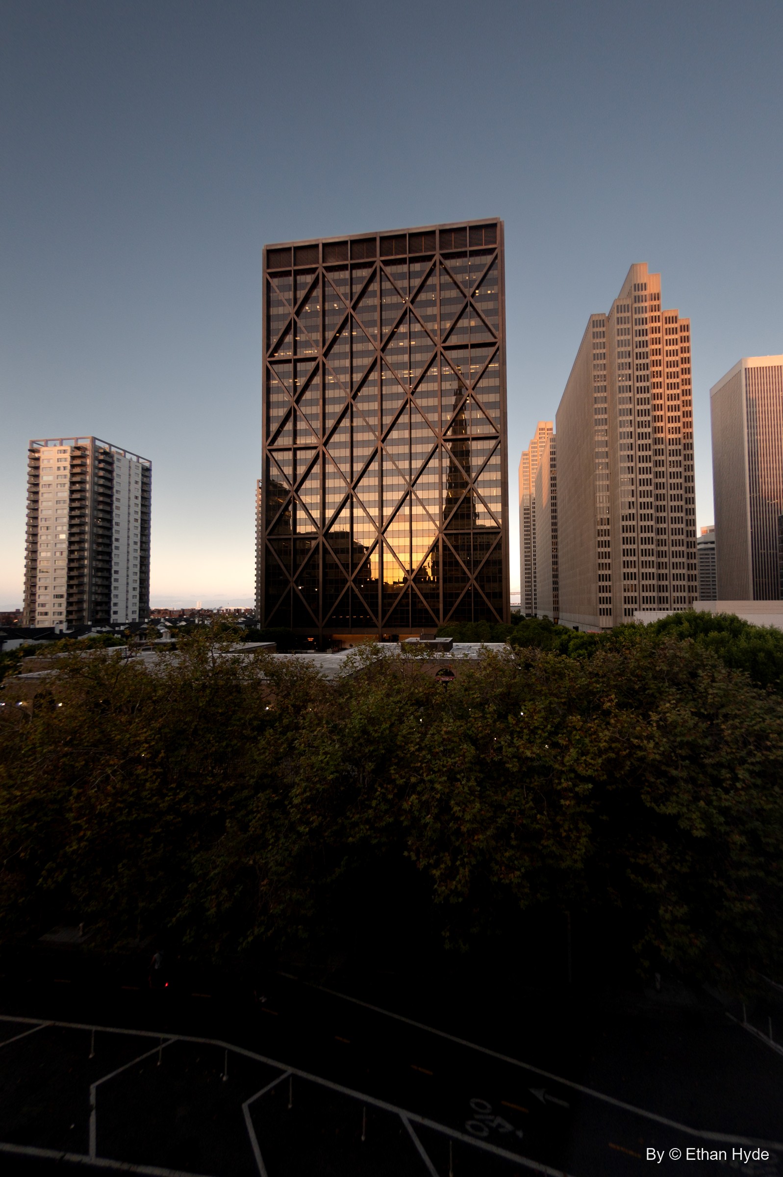 A dark grid-faced office tower catches orange dusk light between pale high-rises, above a band of treetops.