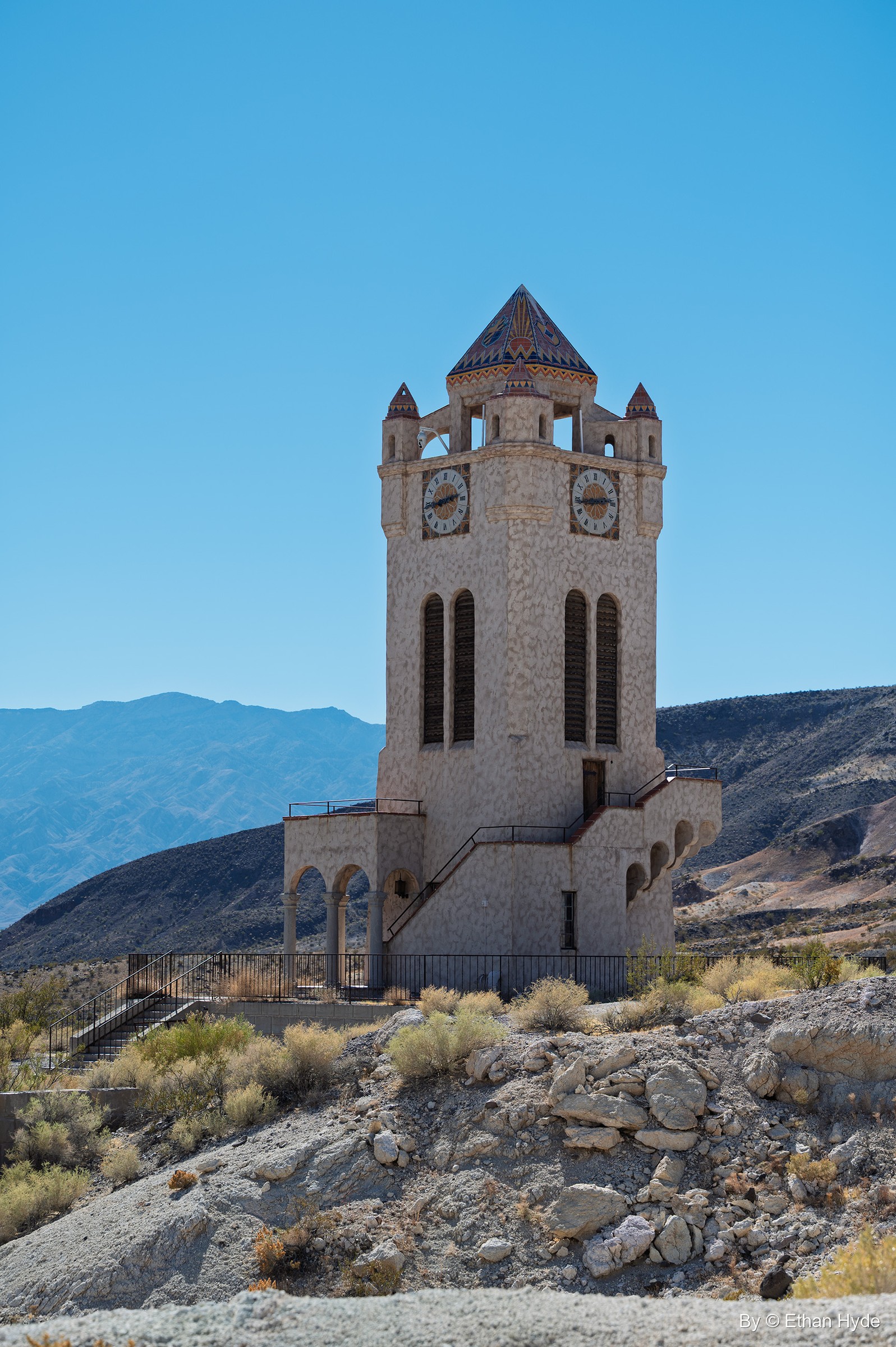The Chimes Tower at Scotty's Castle seen from below on a rocky slope, with distant desert mountains under a clear sky.