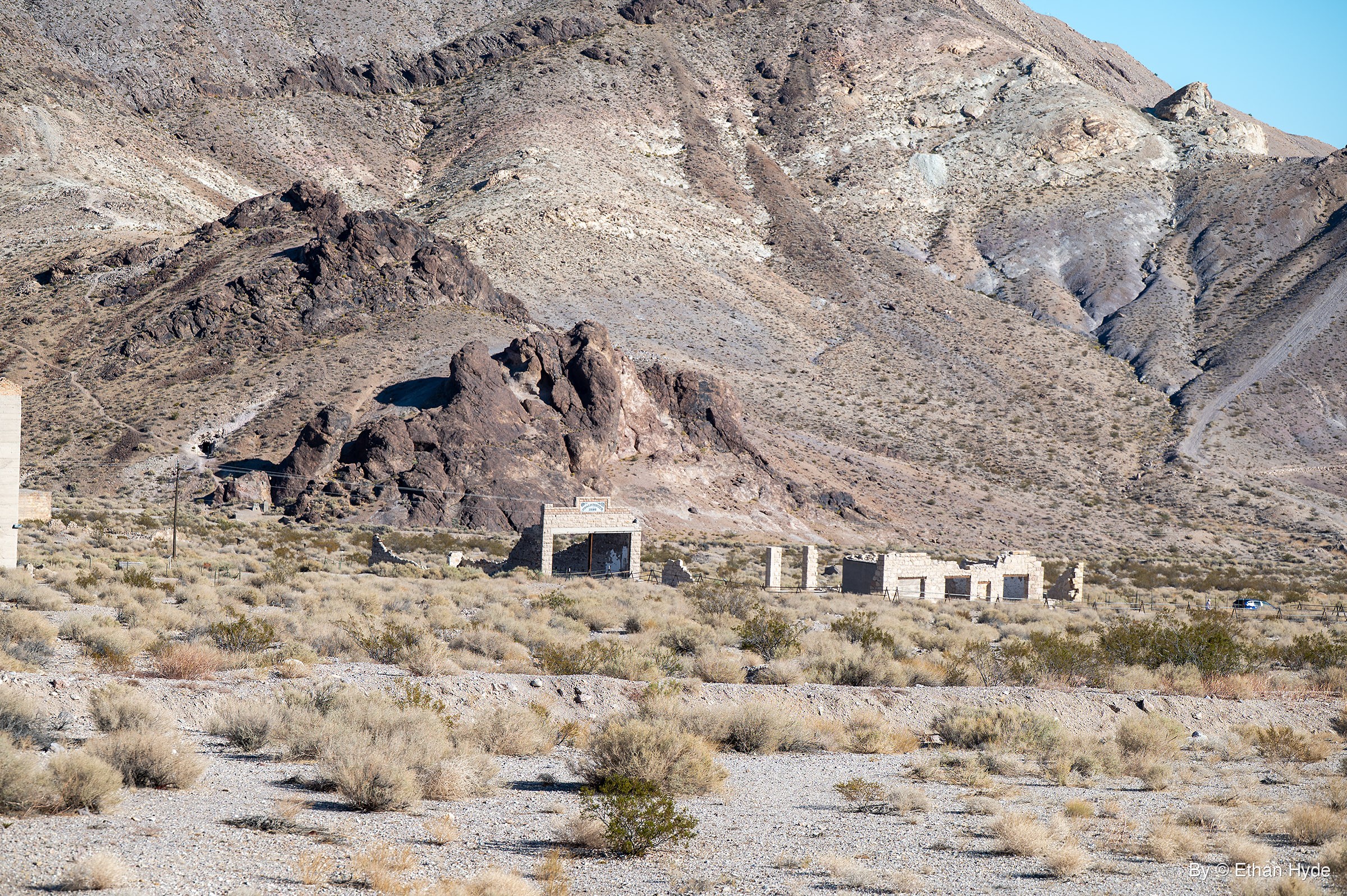 Roofless stone building shells and a freestanding facade stand in dry brush beneath a dark rock outcrop and a steep desert mountainside.