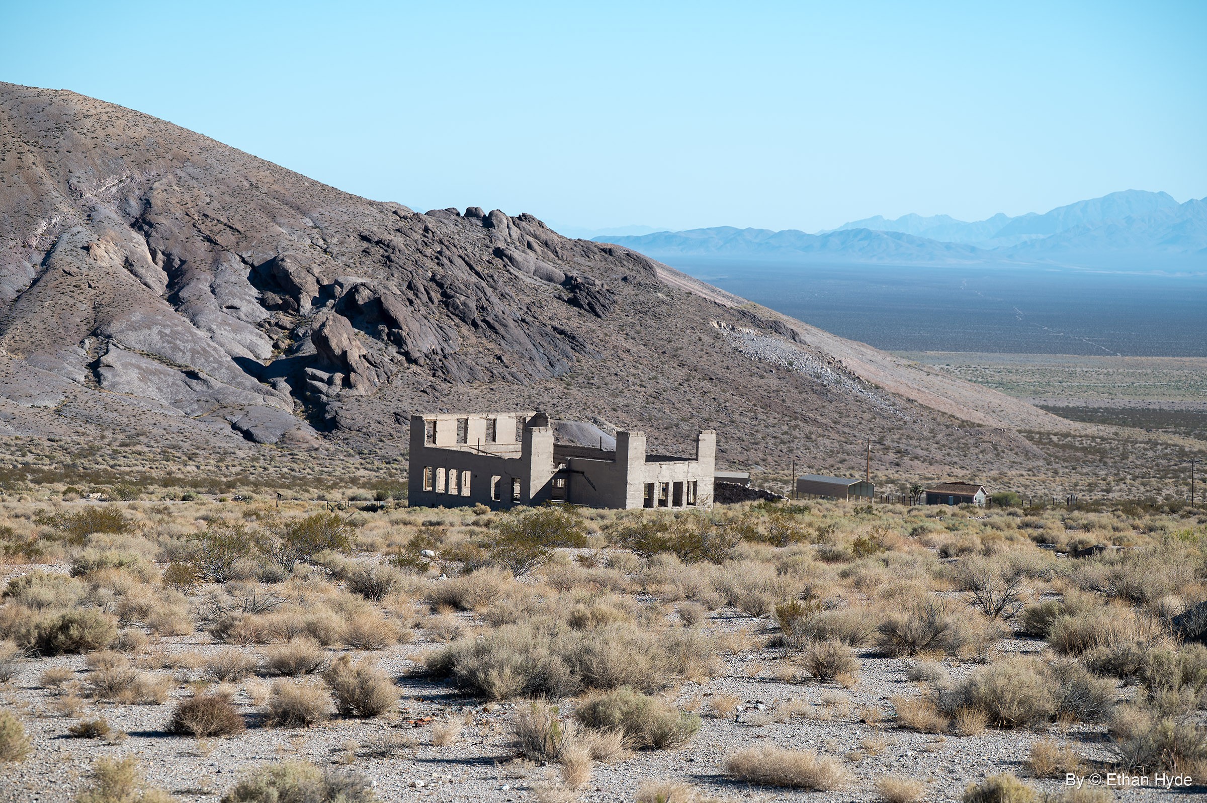 A roofless two-story concrete ruin with empty window openings stands in desert scrub below a rocky hill, with a wide basin and hazy mountains beyond.