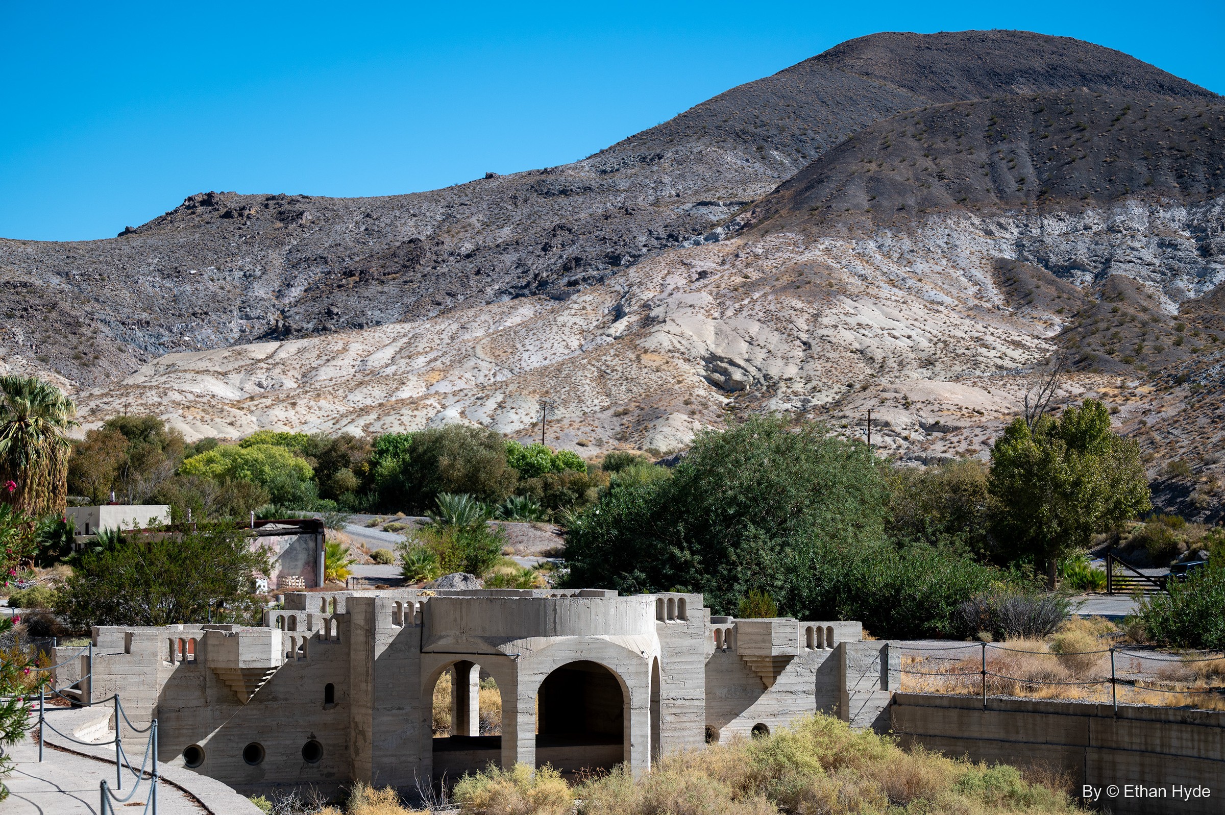 Crenellated walls and arched openings around the unfinished pool area at Scotty's Castle, with oasis trees and banded hills behind.